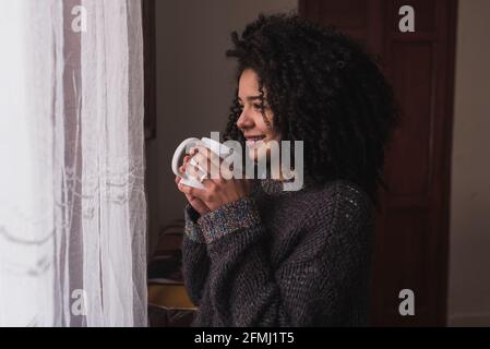 Vue latérale d'une charmante femme ethnique avec des cheveux afro noirs se tenir debout avec une tasse de boisson à la maison et regarder dehors de la fenêtre Banque D'Images