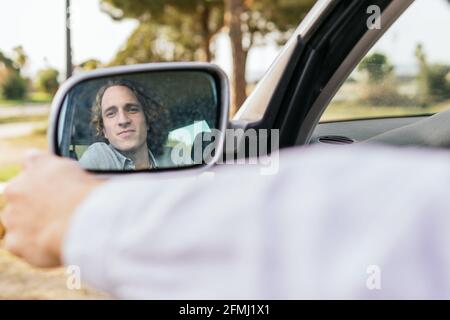 Jeune homme souriant, le conducteur se reflète dans le rétroviseur latéral de voiture garée sur la prairie verte pendant le voyage d'été à travers campagne Banque D'Images