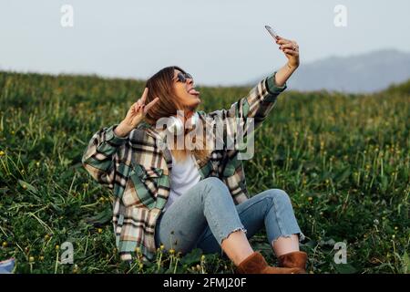 Jeune femme en lunettes de soleil avec casque montrant le geste de paix et en tirant la langue vers l'extérieur tout en prenant autoportrait sur le téléphone portable et assis sur la prairie Banque D'Images