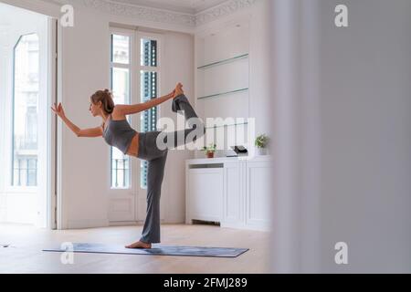 Vue latérale sur l'équilibre paisible des femmes à Natarajasana pendant que vous vous tenez debout sur tapis et pratiquer le yoga dans la chambre à la maison Banque D'Images