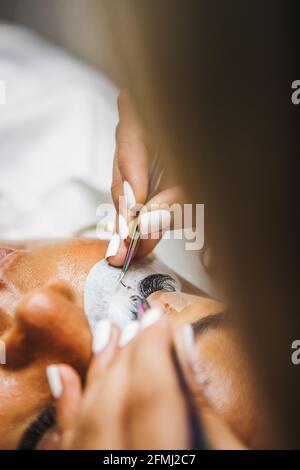 Rognez un cosmétologue méconnaissable à l'aide d'une pince à épiler en appliquant de faux cils pour l'extension à l'œil du client ethnique dans le salon Banque D'Images