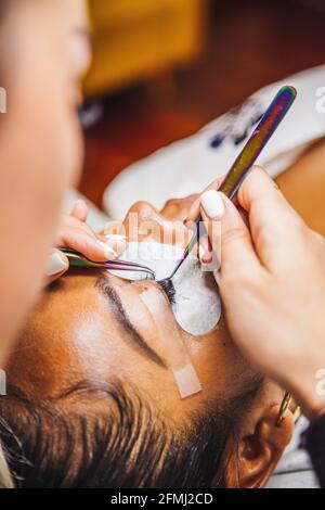 Grand angle de récolte cosmétologue méconnaissable avec des pinces à épiler en appliquant de faux cils pour extension sur l'oeil de client ethnique en salon Banque D'Images