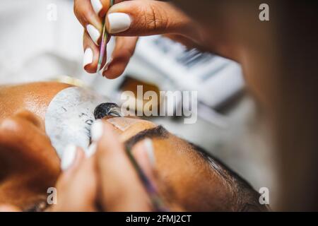 Grand angle de récolte cosmétologue méconnaissable avec des pinces à épiler en appliquant de faux cils pour extension sur l'oeil de client ethnique en salon Banque D'Images