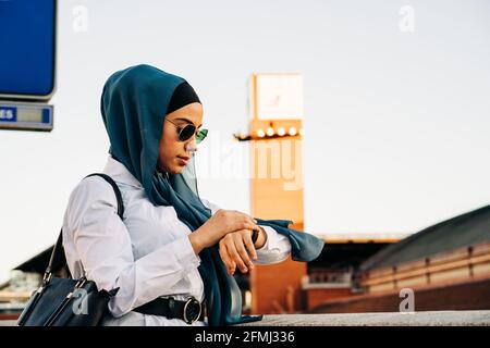 Vue latérale de la femme musulmane dans le hijab debout sur la plate-forme à la gare et vérifier l'heure sur la montre-bracelet en attendant pour le train Banque D'Images