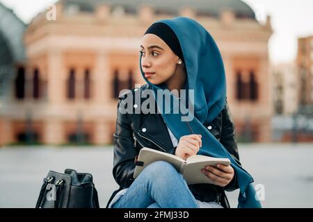 Femme musulmane réfléchie dans le hijab écrivant dans un journal pendant qu'elle est assise sur la rue de la ville et vue loin Banque D'Images