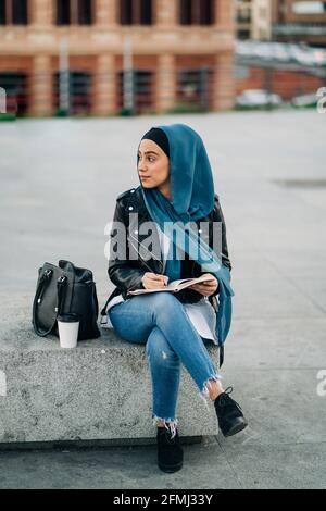 Femme musulmane réfléchie dans le hijab écrivant dans un journal pendant qu'elle est assise sur la rue de la ville et vue loin Banque D'Images
