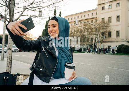 Une femme ethnique gaie portant un foulard traditionnel assis dans la rue boisson à emporter et autoportrait sur smartphone Banque D'Images