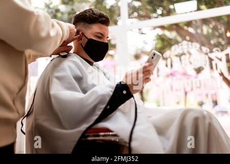 De dessous de Crop stylist avec la tondeuse coupant les cheveux de homme en tissu masque facial avec téléphone cellulaire dans le barbershop Banque D'Images