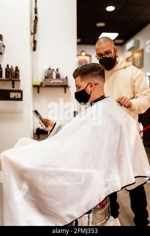 Vue latérale du Crop stylist avec tondeuse coupant les cheveux homme en tissu masque facial avec téléphone cellulaire dans le barbershop Banque D'Images