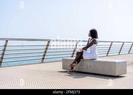 Vue latérale sur toute la longueur afro-américaine tendance dans élégant robe blanche assise avec les jambes croisées sur le banc de pierre promenade de la ville et de profiter Banque D'Images
