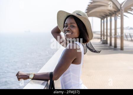 Vue latérale Afro-américaine féminine dans une élégante robe blanche debout sur la promenade de la ville et profiter de la vue sur la mer par temps ensoleillé Banque D'Images