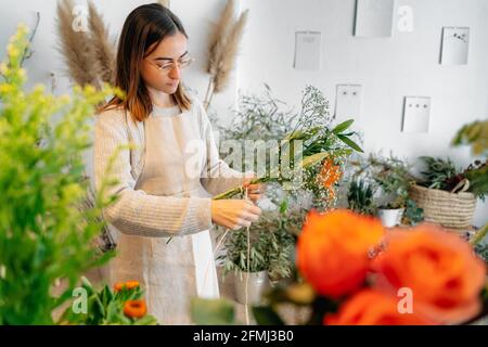 Jeune fleuriste ethnique ciblé en vêtements décontractés et en tablier noue un délicat bouquet de lys orange et de gitsophila blanche boutique de fleurs Banque D'Images