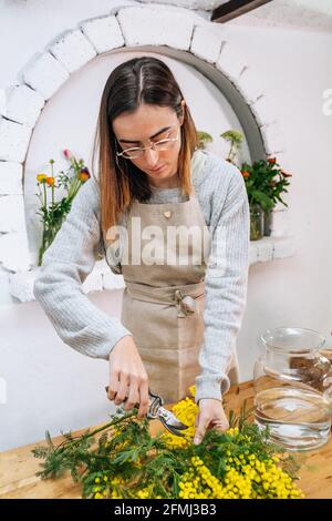 Jeune femme fleuriste dans des vêtements décontractés coupant des tiges de frais fleurs lumineuses avec des cisaille tout en arrangeant le bouquet dans le verre vase Banque D'Images