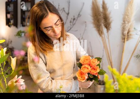 Jeune fleuriste concentré dans un tablier et des lunettes de vue en arrangement parfumé fleurs jaunes dans un vase tout en travaillant dans une boutique florale Banque D'Images