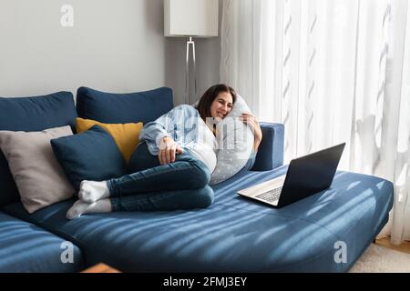 Femme enceinte ravie, couchée sur un canapé confortable et regardant drôle vidéo sur netbook tout en vous relaxant à la maison Banque D'Images