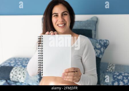 Femme enceinte ravie assise sur un lit confortable à la maison et affichage d'un bloc-notes vierge sur l'appareil photo Banque D'Images