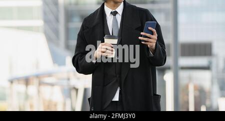 Jeune homme d'affaires ethnique court et peu reconnaissable, bien habillé, avec téléphone portable et une boisson chaude pour aller en ville Banque D'Images