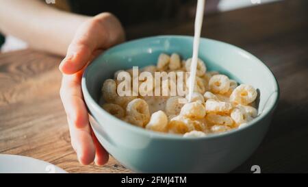 Récoltez une personne méconnaissable en versant du lait dans un bol de maïs délicieux anneaux pour le petit déjeuner sur une table en bois Banque D'Images