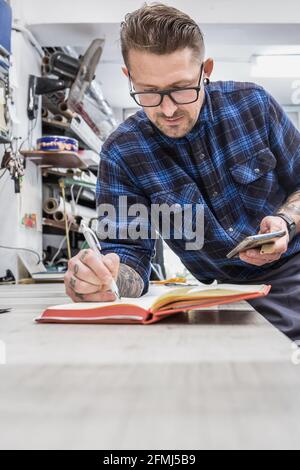 Homme barbu concentré artisan debout à l'établi et à l'écriture bloc-notes lors de l'utilisation du smartphone dans workshop Banque D'Images