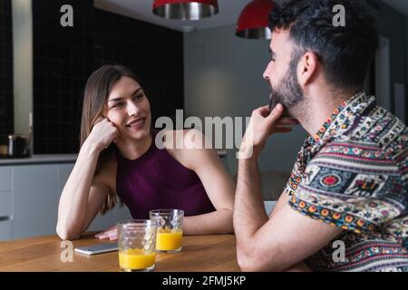 Jeune couple positif en vêtements décontractés ayant la conversation et appréciant jus d'orange frais tout en étant assis à la table à manger dans la cuisine Banque D'Images