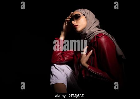 Vue latérale une femme islamique modish et confiante portant un foulard tendance et des lunettes de soleil qui touchent le visage sur fond noir en studio Banque D'Images