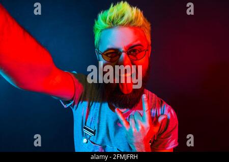 Un blogueur d'hipster enthousiaste avec une barbe et des cheveux verts dans un t-shirt et une combinaison en denim, regardant un appareil photo selfie avec une expression folle, montrant la langue ou Banque D'Images