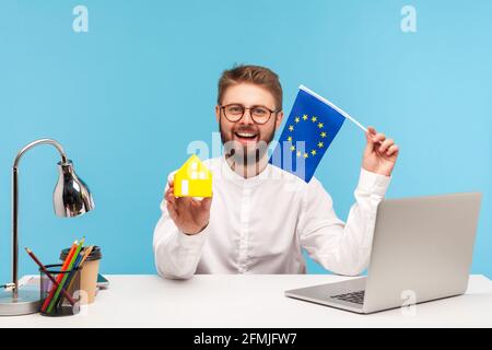 Sourire positif homme agence immobilière travailleur dans les lunettes tenant une maison de jouet en papier et le drapeau de l'union européenne, l'enregistrement des visas et des passeports. Po Banque D'Images