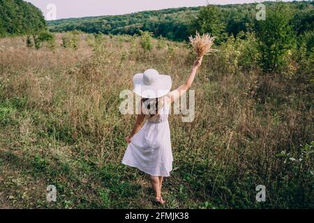 Une femme dans un grand chapeau blanc avec un bouquet marche au milieu de la grande herbe. Fille avec de longs cheveux dans une robe blanche court dans le champ. Une soirée d'été. Banque D'Images