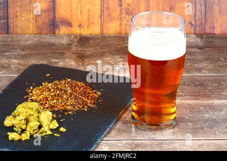 Pinte de verre rempli de bière sur le côté d'un ardoise remplie de houblon et de brasseurs malt d'orge Banque D'Images