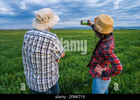 Deux générations de fermiers fiers sont debout dans leur champ d'orge au coucher du soleil. Ils photographient le terrain. Banque D'Images