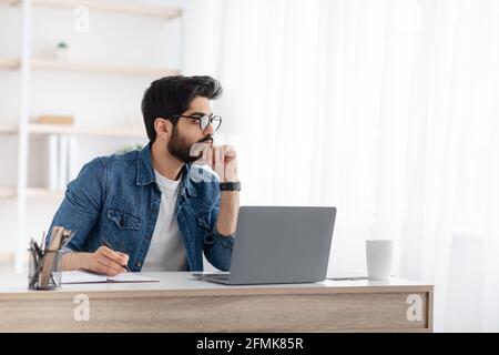 Un indépendant arabe réfléchi qui note des idées sur le bloc-notes, prend des notes sur le lieu de travail à domicile, planifie le programme de travail et le budget, s'assoit au bureau avec un ordinateur portable Banque D'Images