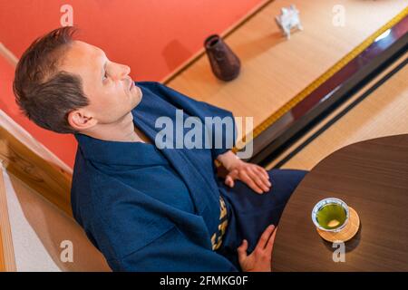 Maison japonaise traditionnelle machiya avec homme dans kimono ou yukata en alcôve rouge buvant du thé vert dans une tasse sur la soucoupe hauteur de la table au-dessus de la vue Banque D'Images