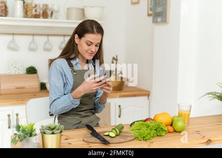 Bonne jeune femme qui boit du jus d'orange et qui regarde à travers les recettes sur son smartphone. Salade végétalienne. Régime alimentaire. Banque D'Images