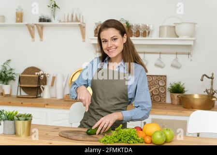 Belle jeune femme souriante préparant une salade de légumes à la maison dans la cuisine moderne loft, trancher un concombre, copyspace et regarder dans l'appareil photo. Banque D'Images