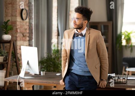 Jeune homme de course mixte sérieux en lunettes debout au bureau table avec ordinateur moderne et vue de côté Banque D'Images