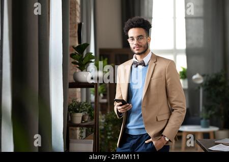 Portrait du jeune homme noir souriant dans une veste élégante et lunettes debout avec la main dans la poche du pantalon et utilisant le gadget dans un bureau moderne et confortable Banque D'Images
