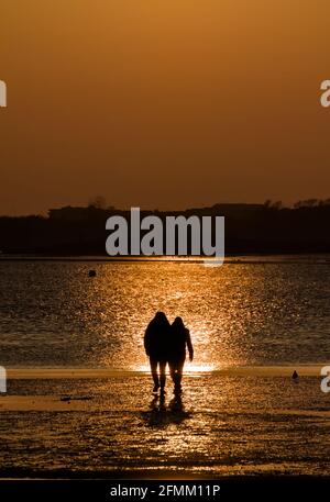 Deux personnes, couple de marche à travers la plage tenant les mains dans le soleil de paramètre au coucher du soleil avec des réflexions dans l'eau, Mudeford Quay UK Banque D'Images