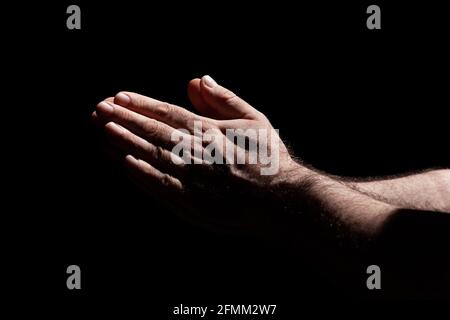 Les mains mâles se replient dans la prière au soleil avec des ombres dures, entourées de ténèbres Banque D'Images