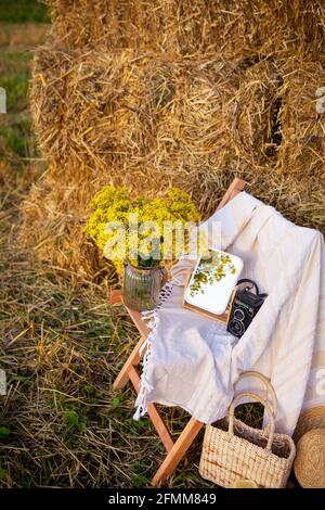 Pique-niquez dans le champ près des balles de paille. Le soleil couchant. Style rustique - chaise en bois, écossais, bouquet de fleurs, miroir, chapeau, appareil photo. Date romantique Banque D'Images