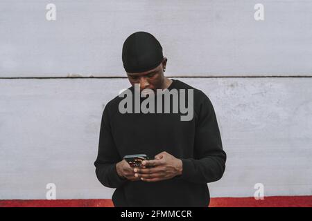 Homme espagnol noir dans des vêtements décontractés noirs et faire-chiffon à l'aide son téléphone contre un mur blanc Banque D'Images