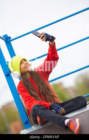 La photo verticale d'une fille de dix ans dans un casque blanc prend un selfie sur un smartphone pour l'envoyer à des amis. Autodétermination et culture de la jeunesse conc Banque D'Images