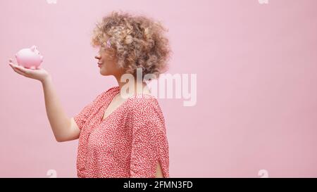 Jeune femme caucasienne aux cheveux bouclés mettant des pièces de monnaie dans la banque de piggy. Concept d'épargne, d'investissement et de richesse croissante. Photo de haute qualité Banque D'Images