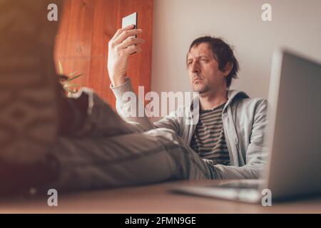 Travailleur indépendant inactif faisant des photos de selfie avec un smartphone au bureau, avec ses pieds sur le bureau, gros plan avec une mise au point sélective Banque D'Images