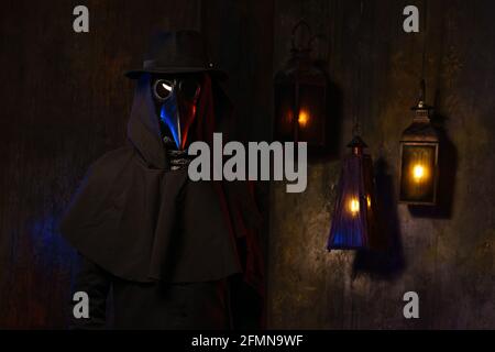 Portrait d'un médecin de la peste dans un masque en cuir noir, chapeau et cagoule, isolé contre un mur sombre avec des lanternes. Stylisation d'un costume historique, d'une image créepy, du concept d'une épidémie. Banque D'Images