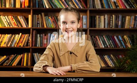 Belle école modèle pose sourire gaiement tout en s'asseyant à nouveau bureau en bois brun avec livres colorés sur des étagères et vert usine de pot Banque D'Images