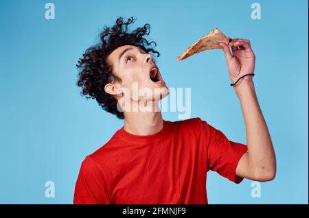 homme avec un morceau de pizza dans sa main un adolescent de fond bleu dans un t-shirt rouge cheveux bouclés Banque D'Images