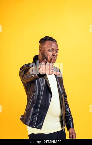 Homme noir confiant portant une veste en cuir noir posant en studio et regardant l'appareil photo sur fond jaune. Banque D'Images