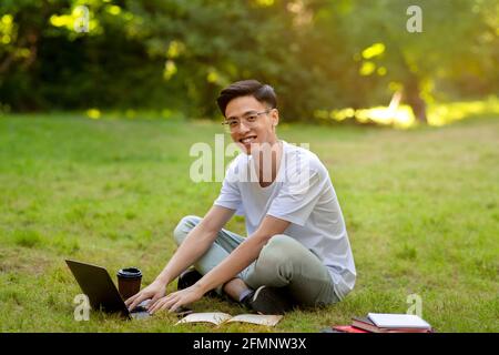 Jeune homme asiatique souriant assis sur l'herbe avec ordinateur, étudiant à l'extérieur Banque D'Images