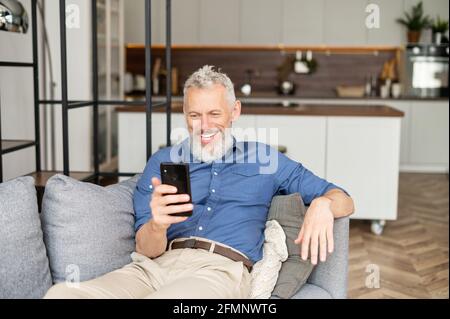 Hilarant charismatique homme mature dans une chemise décontractée utilisant un smartphone allongé sur le canapé à la maison, beau barbu homme défilant des flux d'informations, envoyant des messages, à l'aide d'une application mobile Banque D'Images
