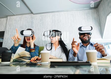 Technologie avancée de réalité virtuelle 3d numérique. Joyeuse surprise trois amis multiraciaux assis dans un café à bouche ouverte et portant des casques VR, essayant un nouveau jeu vidéo ou un projet d'étude Banque D'Images